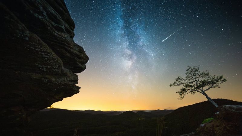  Two meteors, part of the Geminids meteor shower, photographed against the Milky Way with the silhouette of a craggy cliff to one side and a lone tree to the other. Night sky photo taken with a Canon EOS R3 camera and Canon lens by Manuel Dietrich.