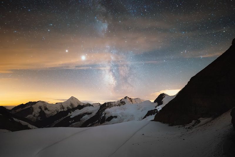 Jupiter and Saturn in conjunction, seen as very bright points of light alongside the Milky Way above snow-covered peaks. Night sky photo taken with a Canon EOS R5 camera and Canon lens by Manuel Dietrich.