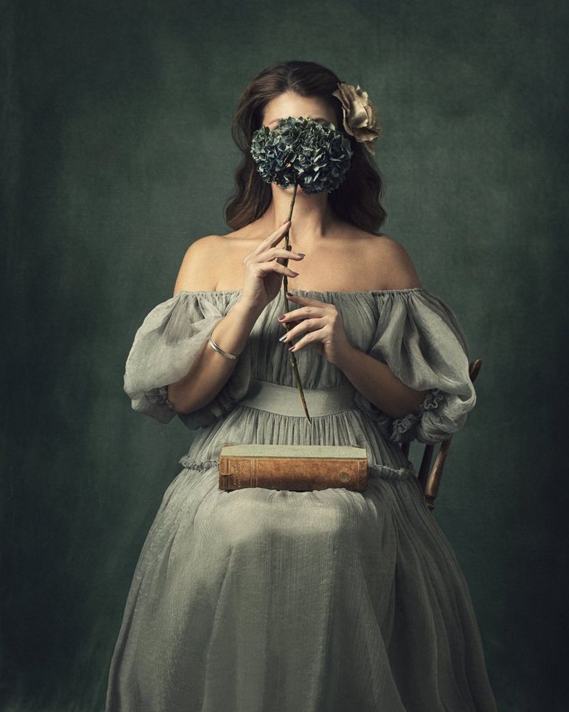 A fine art portrait of a seated woman in grey with a leather-bound book in her lap, holding a large flower in front of her face with both hands. Taken on a Canon EOS 5D Mark III.