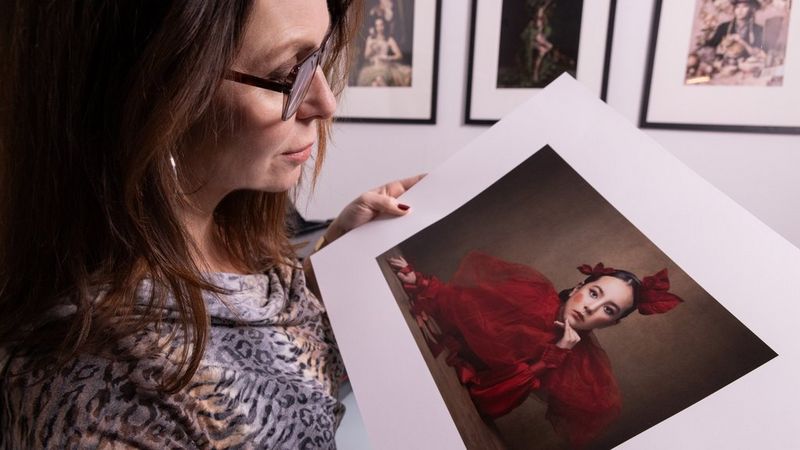 Martina Wärenfeldt holds an A3 photo print of a woman in a deep red outfit, printed on a Canon imagePROGRAF printer. Other framed photo prints hang on a wall behind.