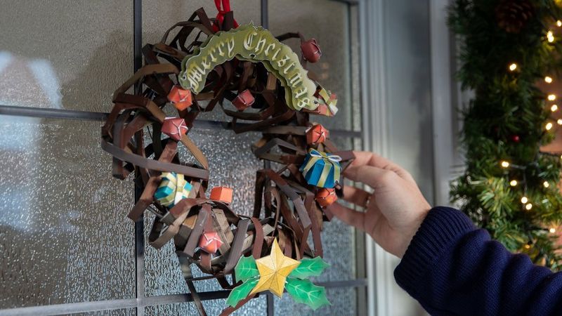 A sumptuously decorated Creative Park papercraft wreath hangs on a front door, with a hand adjusting one of the presents attached to it.