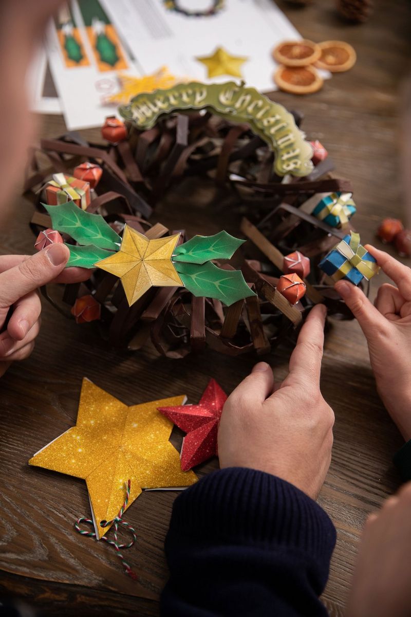 Hands positioning a papercraft gift box on a Creative Park papercraft Christmas wreath.