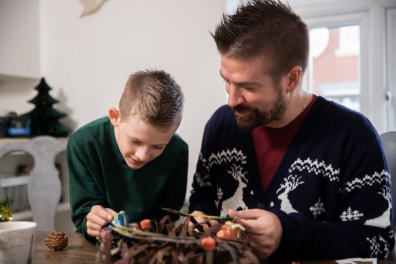 A father and son sit at a dining table leaning over a papercraft wreath they are making together from a Creative Park template.