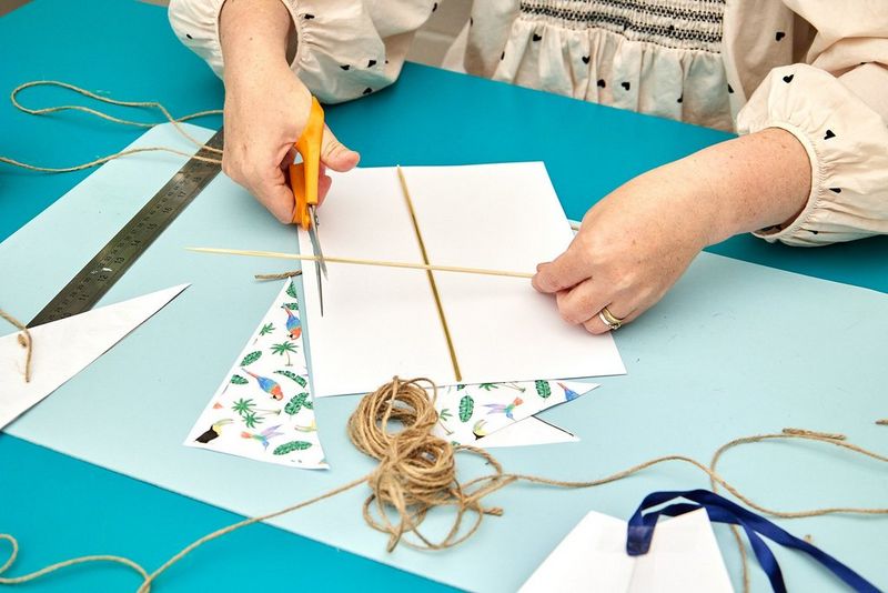 Twee handen snijden houten stokjes op maat die de basis van een zelfgemaakte papieren vlieger vormen.