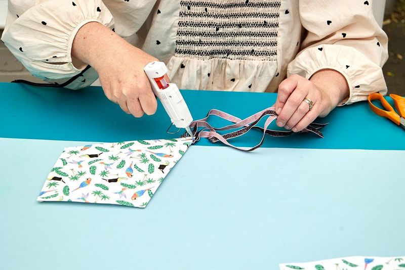 Twee handen houden een lijmpistool vast en plakken daarmee roze en paarse linten vast aan de onderkant van een papieren vlieger.