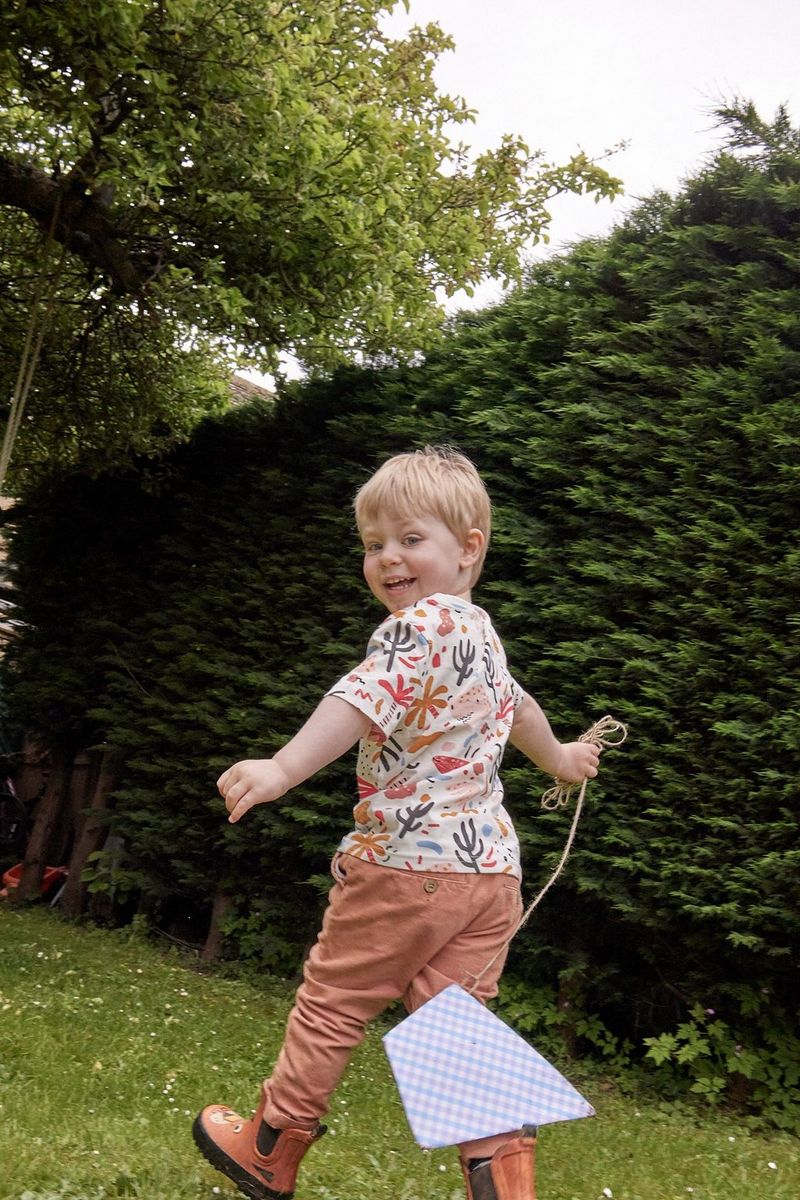 Een jongetje loopt over een gazon en trekt een papieren vlieger aan een touw achter zich aan. Zijn breed lachende gezicht is naar de camera gericht.