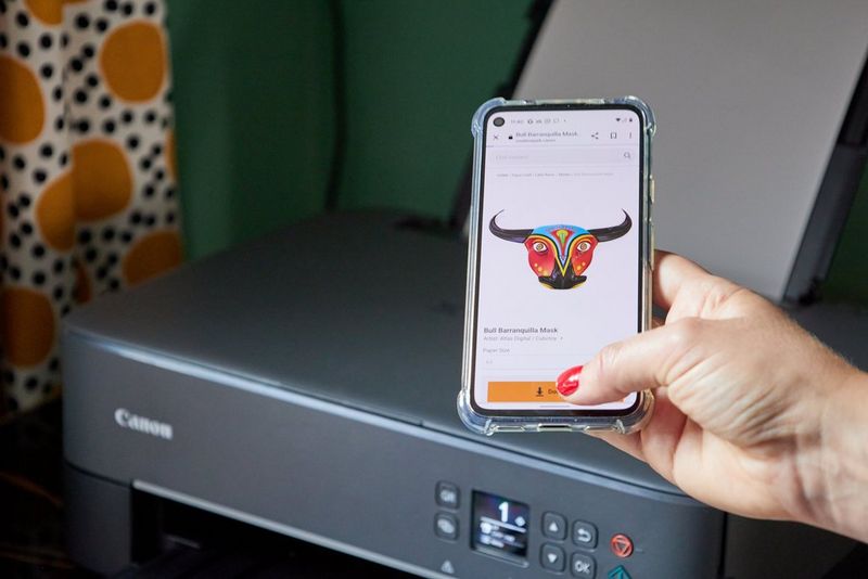 A hand holds a smartphone above a Canon PIXMA printer, a bull mask template from the Canon Creative Park app visible on the screen.