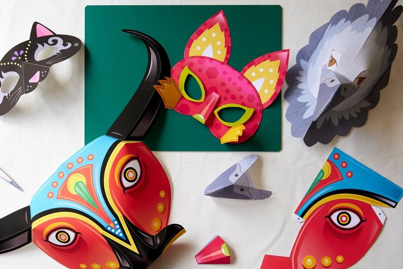 Assembled cat, fox, eagle and bull masks are placed on a table next to a cutting board and other folded pieces of paper.