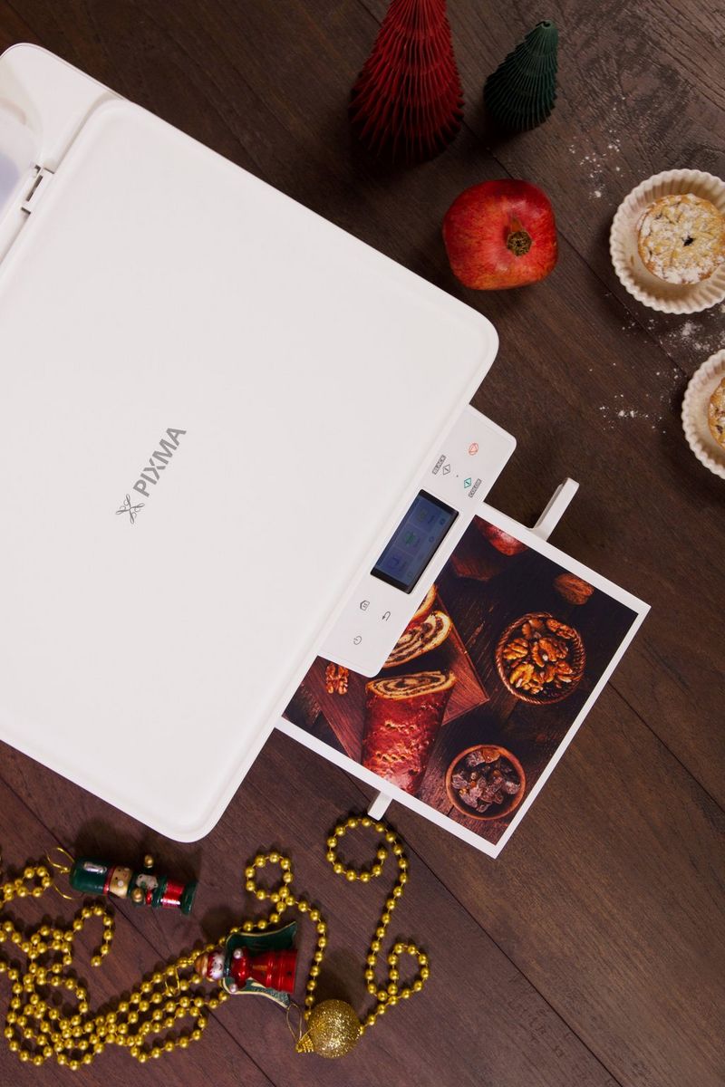 A print of food emerges from the output tray of a Canon PIXMA printer with festive decorations and foods alongside, all viewed from above.
