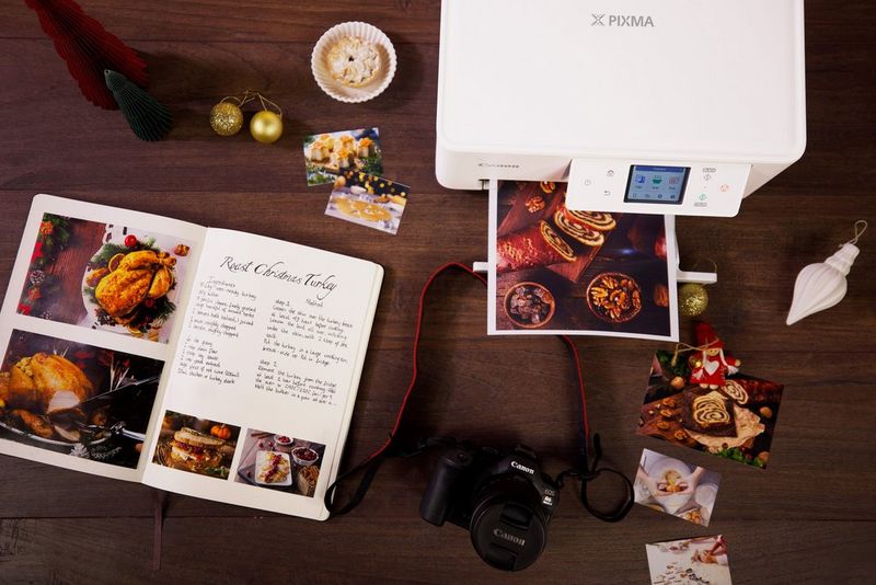 A bespoke recipe book sits on a wooden table alongside small pictures of food, Christmas decorations, a Canon camera, and a Canon PIXMA printer with a large print of food in its output tray.