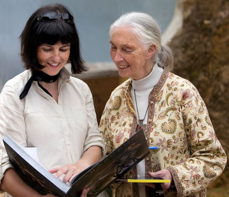 Photographer Marina Cano holds a large book open in her hands, showing it to naturalist Jane Goodall, who stands at her side.
