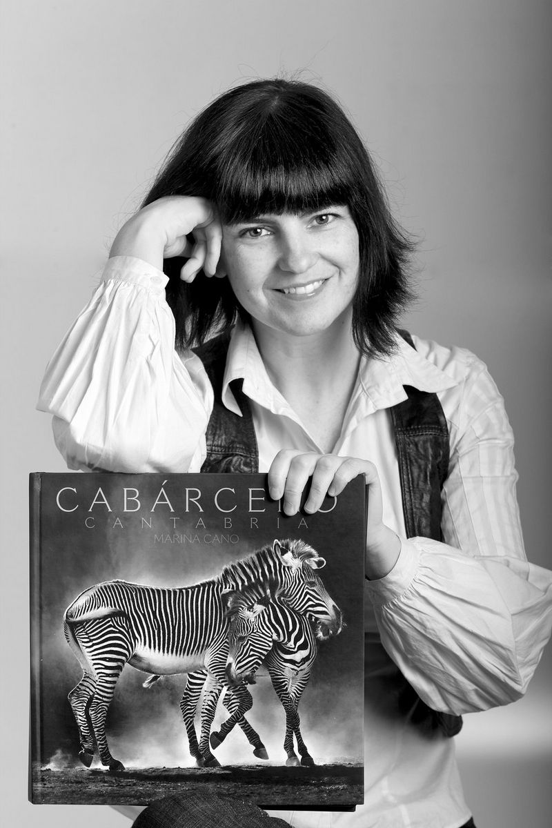 Photographer Marina Cano smiles at the camera, with one elbow leaning on a large book with a black and white photo of zebras on the cover.