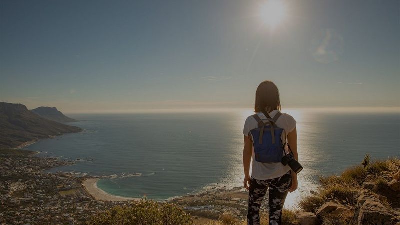 A casually-dressed woman, with a backpack and a Canon EOS RP camera over one shoulder, looks out to sea at a sunset.
