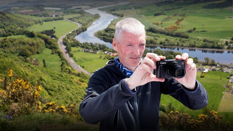 Alan Rowan stands on the side of a hill, with yellow gorse bushes, grassy fields and a river in the valley behind him. He holds a compact camera to take a photo of the natural view.