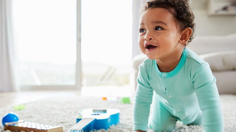 An older baby crawls on a white rug, with a toy guitar next to it.
