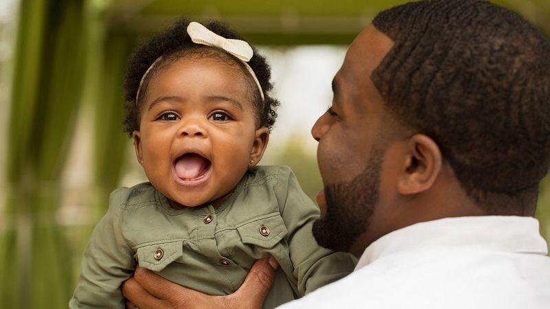 A baby wearing a headband faces the camera. Her dad holds her up above his shoulder in one hand, facing away from us as he looks at her.