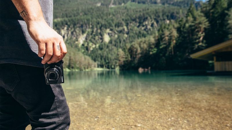 A man, shot from the shoulders down, stands by a lake holding a Canon PowerShot G5 X Mark II in his hand.