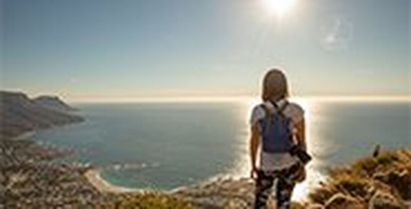 A casually dressed woman, with a backpack and a Canon EOS RP camera over one shoulder, looks out to sea at a sunset.