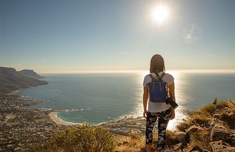 A casually dressed woman, with a backpack and a Canon EOS RP camera over one shoulder, looks out to sea at a sunset.