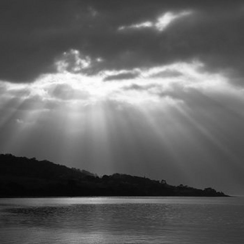 A black and white picture of a coastline with a burst of sunlight above.