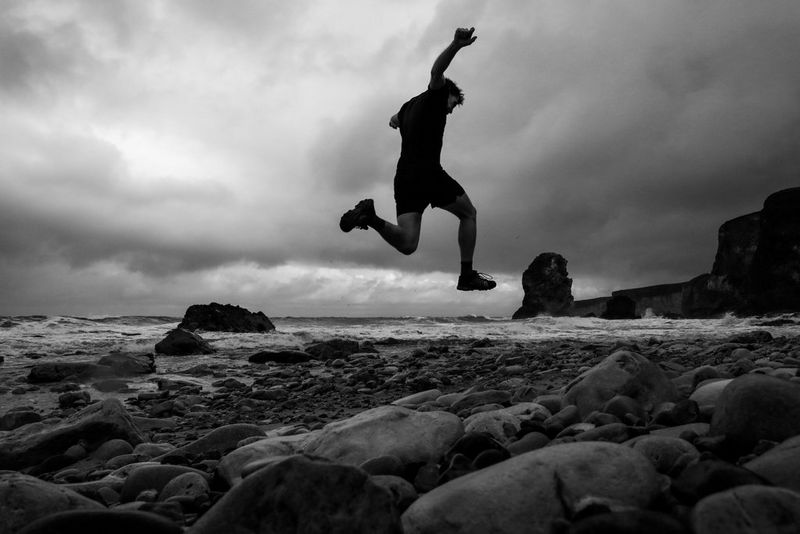 A low-angle shot shows a man in the air as he runs on a rocky beach.