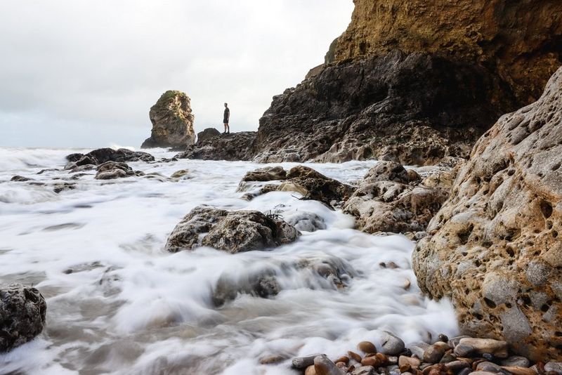 A man stands on rocks by the coast, with the moving waves blurred and softened by a long exposure.