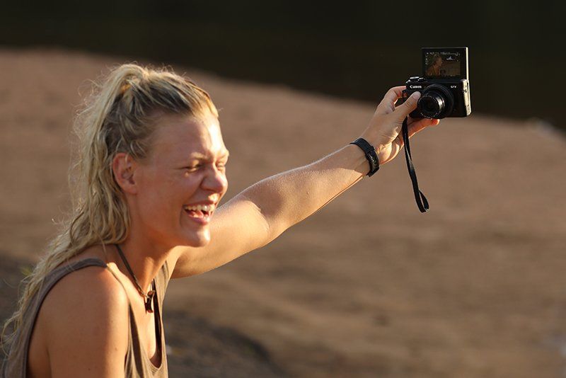 Ness Knight sits on a sandy riverbank filming a vlog with a Canon PowerShot G7 X Mark II.