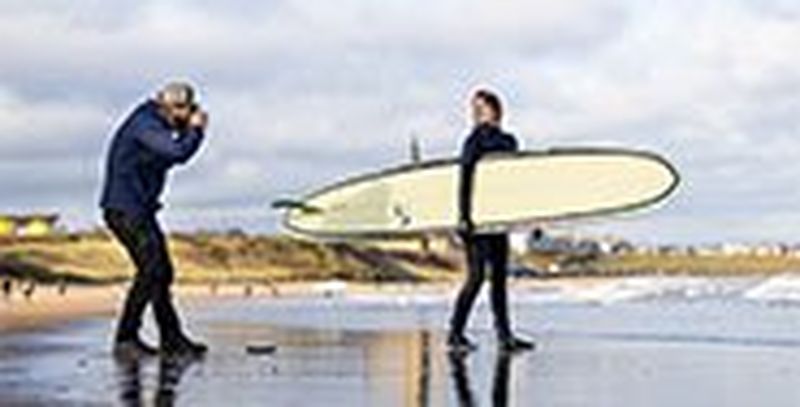 Photographer Tom Bing on a beach taking a picture of a surfer holding her board.