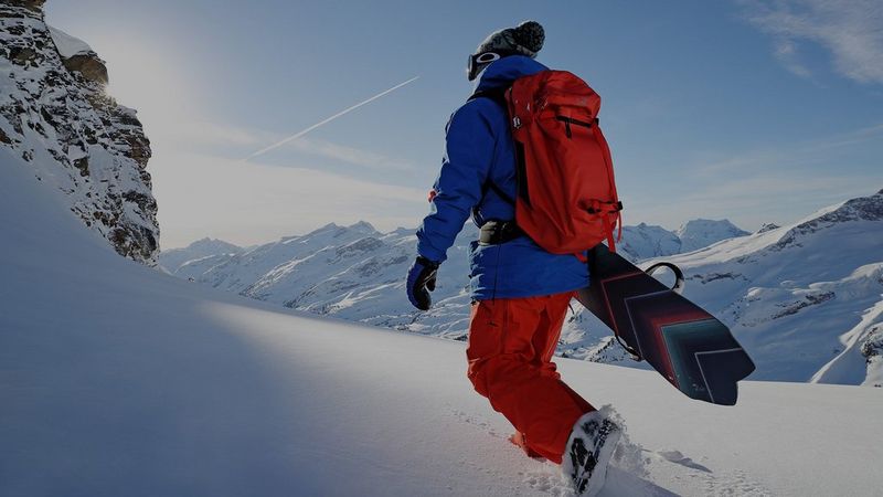 A snowboarder walks through the snow with his snowboard under his arm, away from the camera.