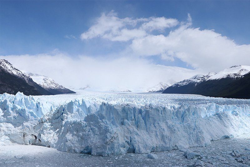 An imposing wall of snow with mountains and blue sky in the background.
