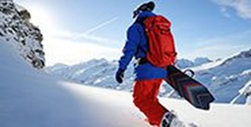 A snowboarder in red trousers walks through the snow, carrying his board, with his back to the photographer.