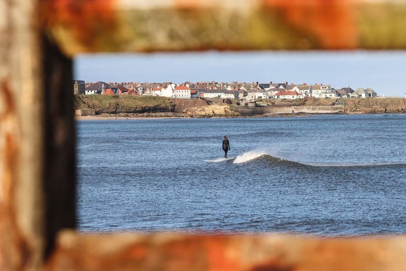 A surfer catching a wave is framed in the bars of a fence in the foreground.