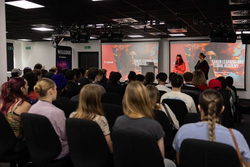 An audience of young people watch two presenters on stage. Behind them are two screens with ‘Canon Learning Day, Global Academy’ printed on them.