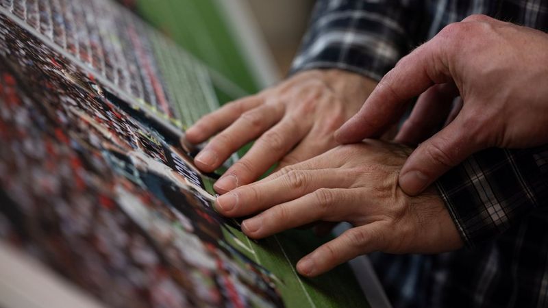 An older woman with short hair, earrings and glasses interacts with a World Unseen exhibit. In front of her are large black and white images of hands. Her hands touch similar black and white images which are mounted on a plinth below.