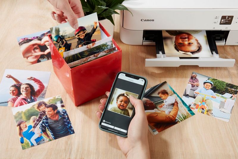 A person's hand holding a phone with a baby photo on the screen, while another hand selects an image from an open Canon Photo Cube. 
