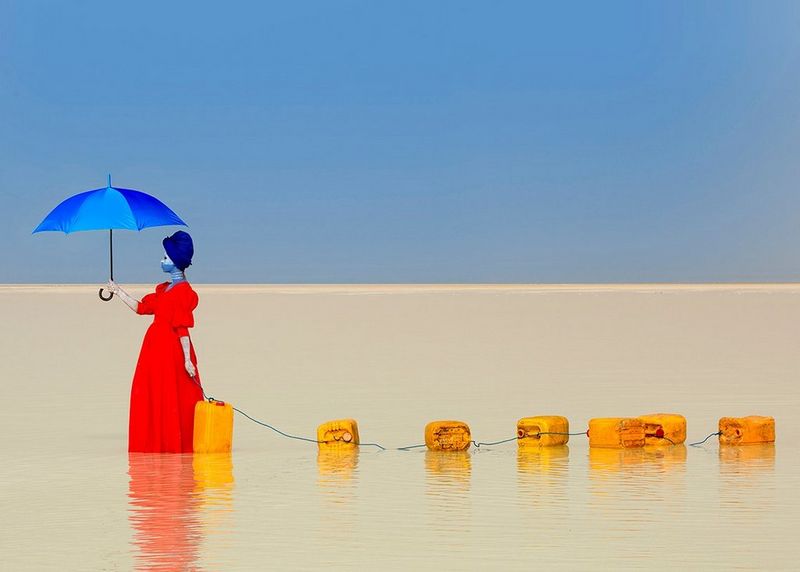A woman in a red dress and holding a blue umbrella walks through shallow water dragging a chain of jerry cans behind her.