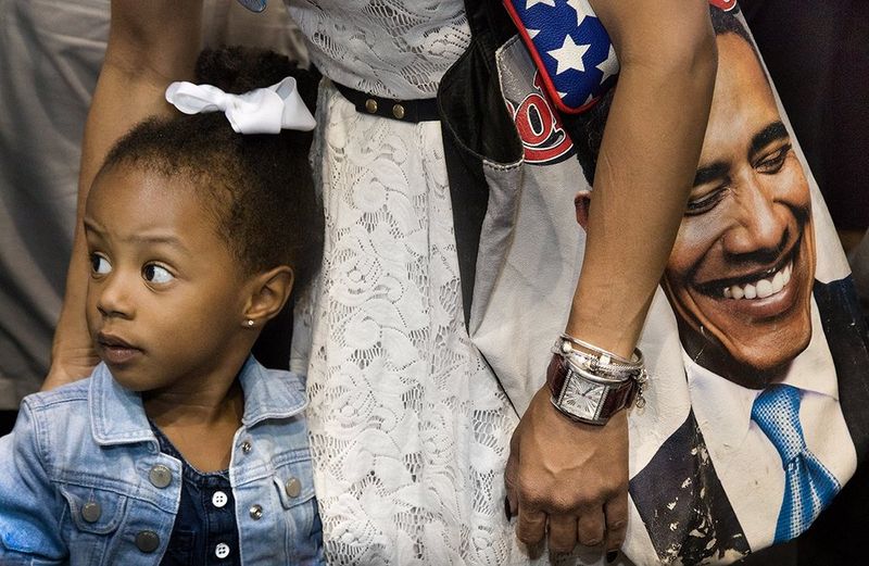 A girl is looking away from her mother, who wears a large Obama bag over her shoulder, which features an image of the President.