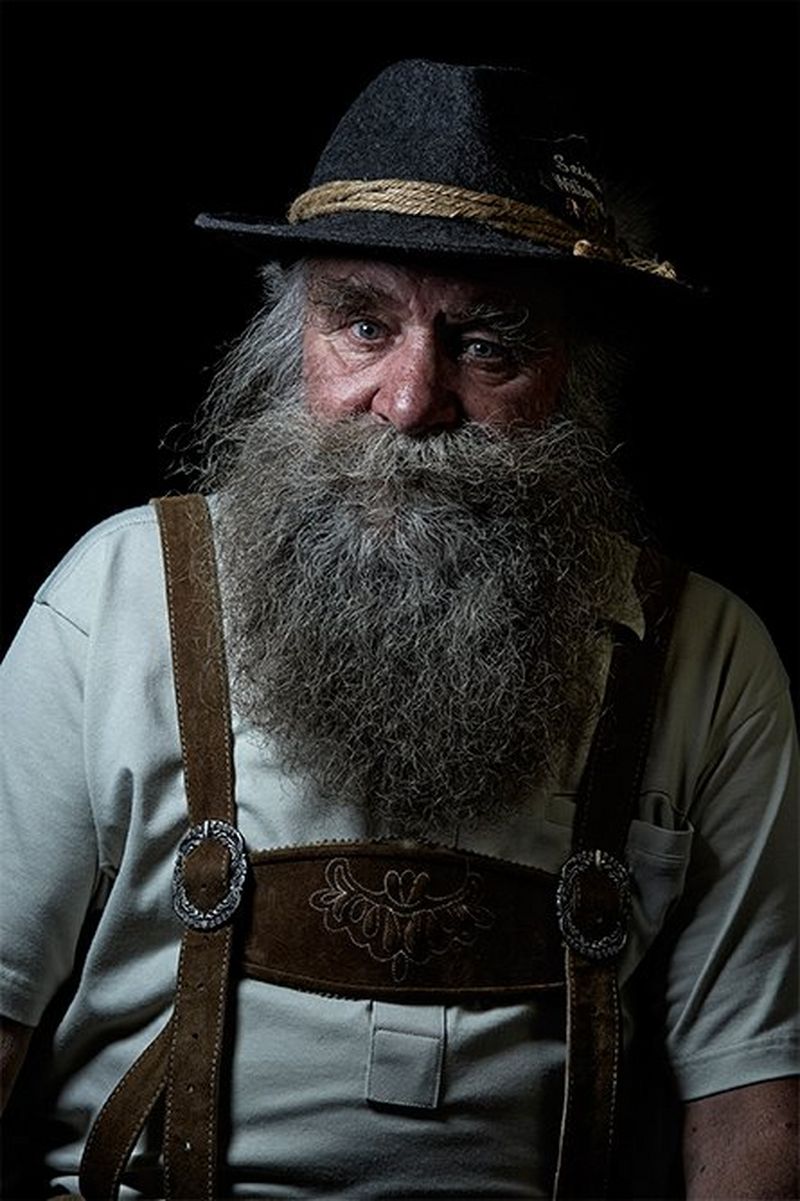 A man with bushy eyebrows and a chest-length grey beard wearing traditional Alpine costume including decoratively-embroidered leather braces