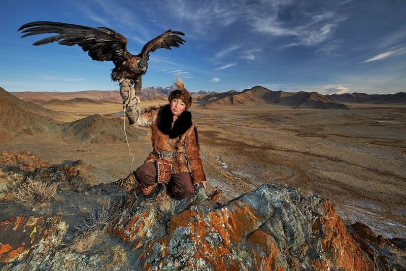 A teenage Mongolian eagle huntress crouches on rocks holding an eagle aloft. Taken by Alessandra Meniconzi on a Canon EOS R.