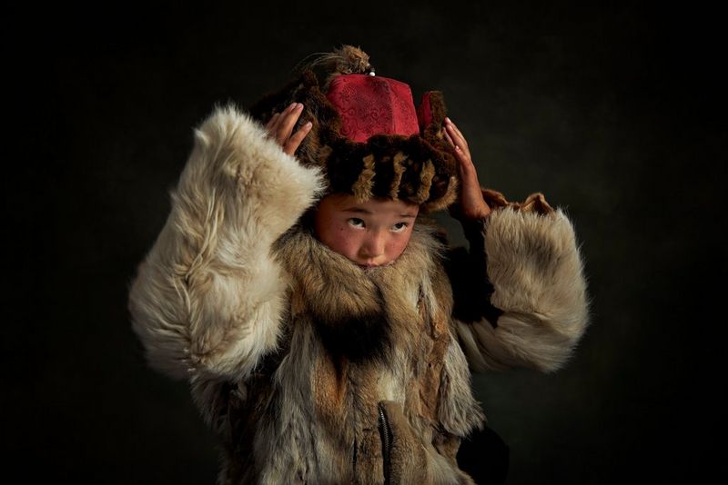 A Mongolian child in a fur coat adjusting her hat with both hands. Taken by Alessandra Meniconzi on a Canon EOS R.