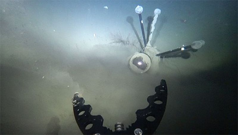 An underwater shot, with the camera's lens and tripod sticking out of the silt just beyond the drone's grabbing arm.