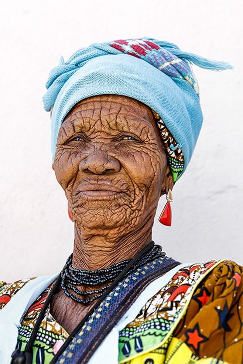 An elderly Namibian woman wearing a colourful patterned dress, dark blue beaded necklace, light blue headscarf, and earrings with large, triangle-shaped red stones.