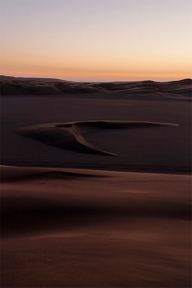 The dunes of the Namibian desert rise to undulating ridges in the distance, with a distinctive dune shaped like a barbed arrowhead in the middle distance.