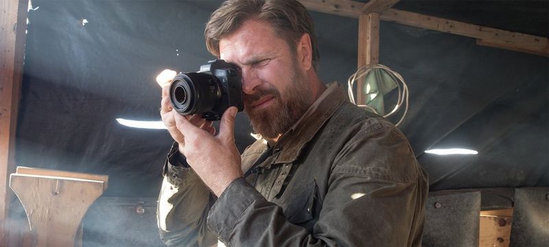Photographer Brent Stirton stands in a structure made of timber and tarpaulin, pointing his Canon EOS R camera at something out of shot.