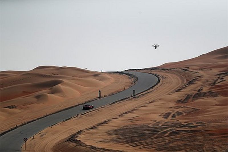 The orange car drives through desert, a large drone filming it from above.
