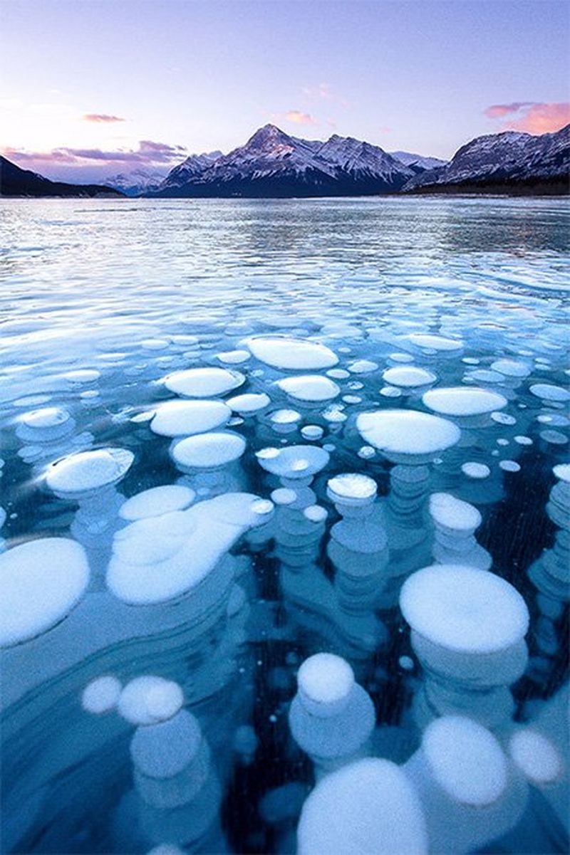 In the foreground, the surface of a frozen lake shows white bubble shapes frozen. In the background are mountains tinged purple-pink by the setting sun.