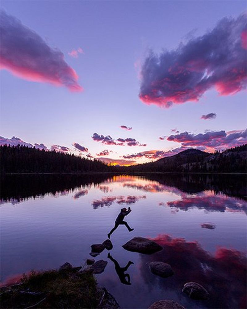 A man leaps between two stepping stones in front of a lake, the sky purple with the setting sun.