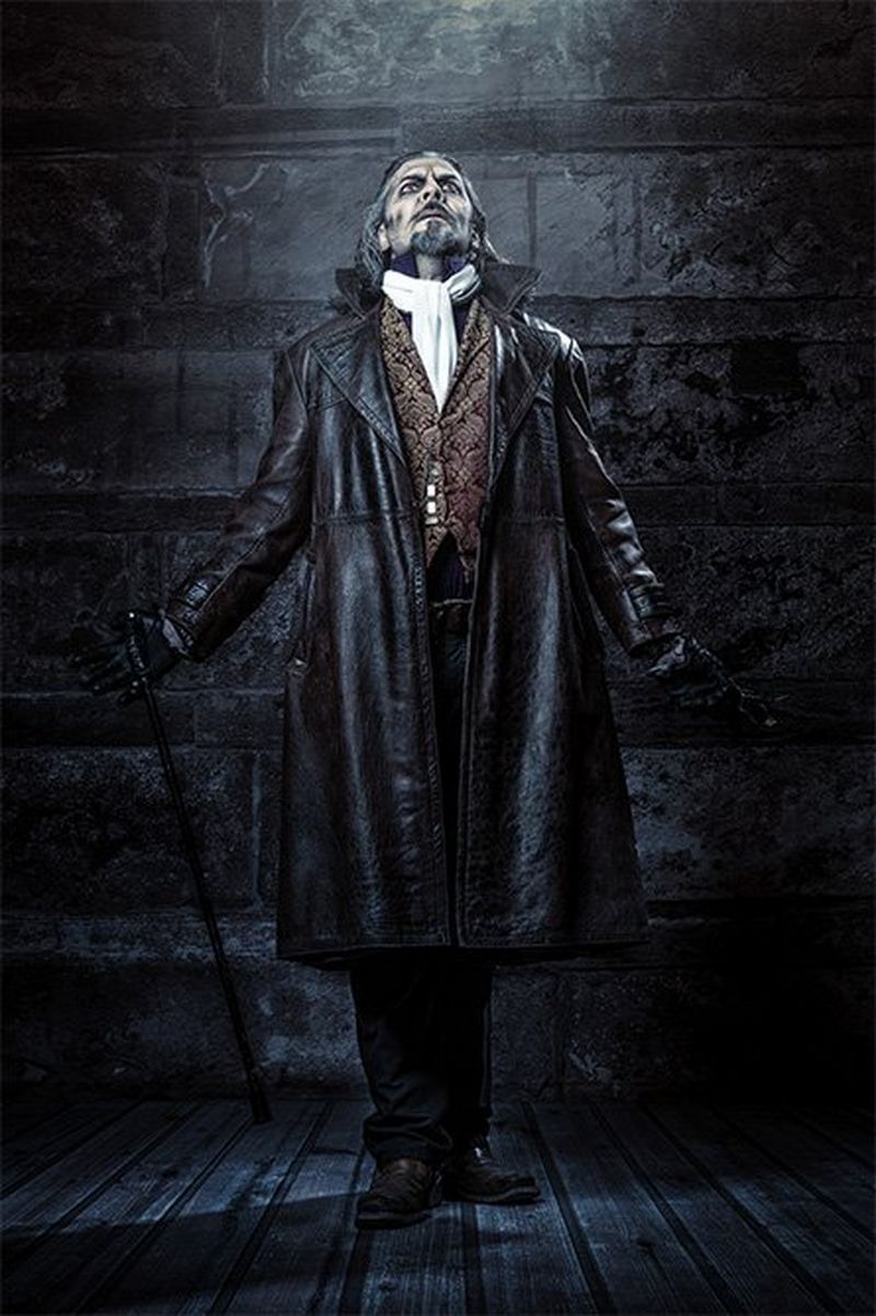 A man is dressed in a black leather cape and carries a cane.