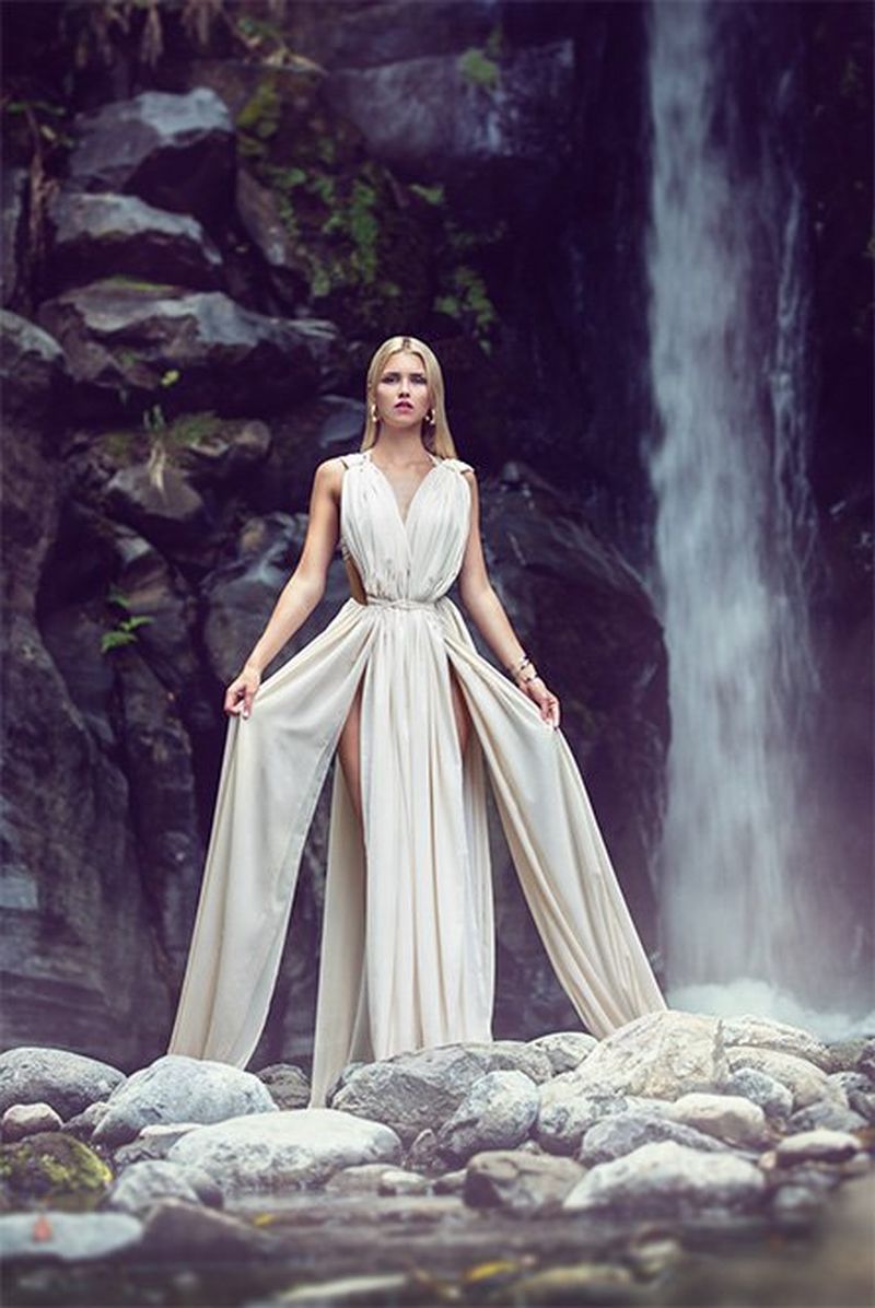 A woman wears a white dress in front of a waterfall.