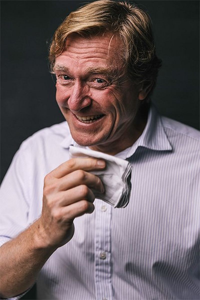 A middle-aged man, smiling broadly, raises a folded handkerchief with one hand.